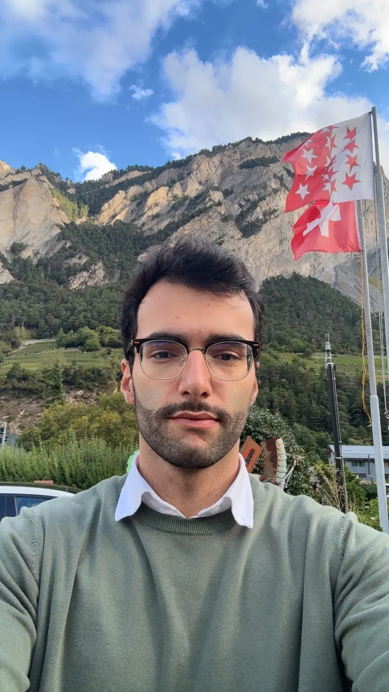 Ali in Switzerland, with mountains and Swiss flags behind him